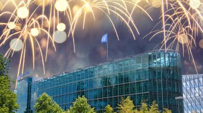 A modern glass office building with a blue flag on the roof, surrounded by trees, with fireworks lighting up the night sky above.