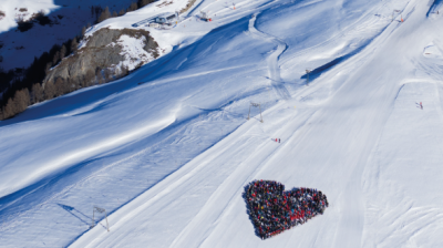 Aerial view of a snowy landscape where a group of people is gathered in the shape of a heart on the snow.