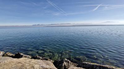 Clear blue lake with visible rocks beneath the water in the foreground, distant mountains on the horizon, and several contrails in the sky.