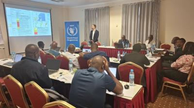 A group of people attend a presentation in a conference room with a World Food Programme banner and projected slides on the wall.