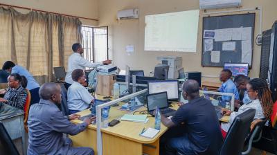 Several people work at computers in an office while one person stands at the front presenting information projected onto a screen.