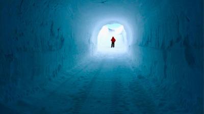 A person wearing a red jacket stands at the end of a tunnel made of snow and ice, illuminated by natural light.