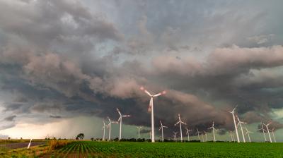 Wind turbines in a field with storm clouds overhead, green crops in the foreground, and dramatic sky in the background.