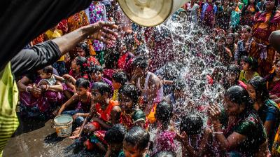 A group of children sit closely together as water is poured over them from a bucket, surrounded by a crowd of onlookers in colorful clothing.