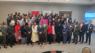 A large group of professionally dressed adults pose together in a conference room, with banners and flags in the background.