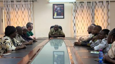 A group of people sit around a conference table, listening to a man seated at the head of the table in an office setting.