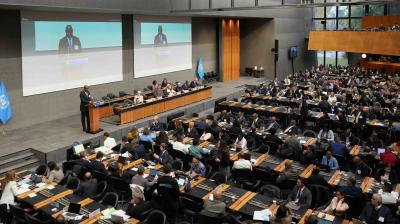 A large conference room with delegates seated at long tables, a speaker at a podium, and two projection screens displaying the speaker at the front.