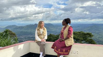 Two women sit on a low wall overlooking a scenic mountain landscape under a cloudy sky. Both wear beige vests; one is in a dress, the other in pants and a jacket.