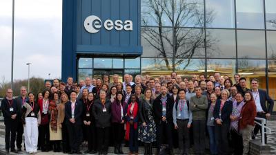 A large group of people stands in front of the ESA building, posing for a group photo. The glass facade reflects trees and sky in the background.