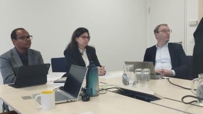 Three people sit at a conference table with laptops, notebooks, a water bottle, and glasses, appearing to attend a meeting in a modern office setting.