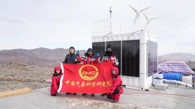 Four people in red jackets hold a red flag with Chinese text and "AMS" logo in front of a solar- and wind-powered research station on a rocky mountain.