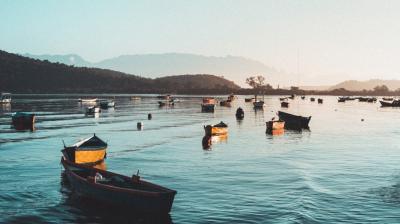 Numerous small boats float on a calm body of water, surrounded by distant hills under a clear sky at either sunrise or sunset.