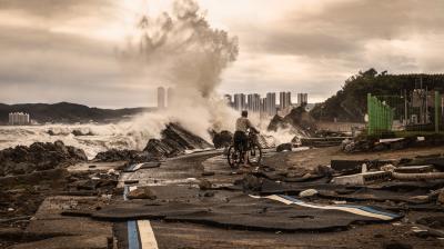 A person with a bicycle stands on a damaged coastal road, watching large waves crash onto rocks; debris and high-rise buildings are visible in the background.