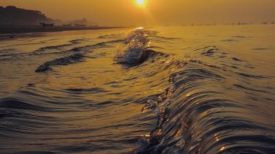 Golden sunlight reflects off gentle ocean waves near the shore at sunrise. Boats and silhouettes are visible in the distance under an orange sky.