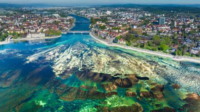 Aerial view of a city with a river flowing into a large lake, showing clear turquoise water, rocky formations, and surrounding urban and green areas.