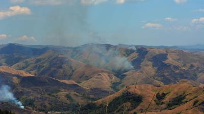 A mountainous landscape with brown, dry hills and scattered smoke rising from multiple areas, under a partly cloudy sky.