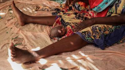 A newborn baby lies on a cloth between an adult woman's legs, with both resting on a patterned fabric floor.