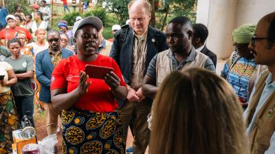 A woman in a red shirt speaks to a group of people gathered outdoors, some wearing badges and vests, while others observe in the background.