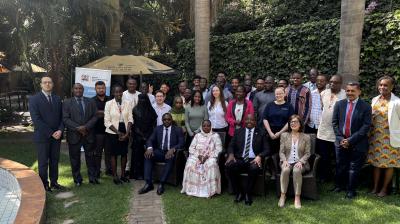 A diverse group of people pose for a formal group photo outdoors, with some seated and others standing, surrounded by greenery and palm trees.