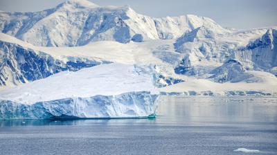 Large iceberg floating in calm water with snow-covered mountains in the background under a cloudy sky.