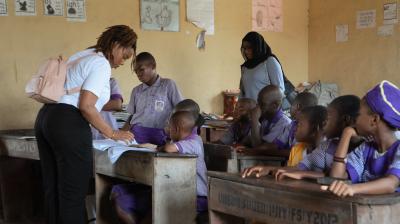 A teacher assists students in a classroom as another adult observes; students are seated at wooden desks wearing purple uniforms. Educational posters are on the walls.