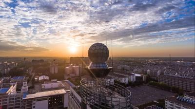 A cityscape at sunset featuring a spherical structure atop a rooftop, with buildings and a cloudy sky in the background.