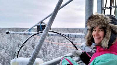 A person in a fur-lined hooded winter coat stands on a metal structure overlooking a snowy forest, smiling at the camera.
