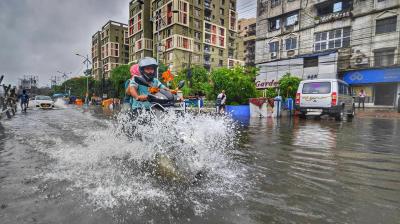 A person rides a motorcycle through a flooded city street, splashing water, with buildings and vehicles in the background on a cloudy day.