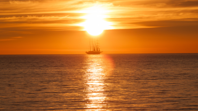 A tall ship sails on calm water at sunset, silhouetted against the bright orange sky with the sun low on the horizon and its reflection on the sea.
