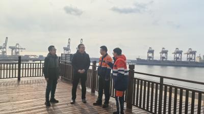 Four people stand on a wooden deck by the water with cranes and industrial structures visible in the background under a cloudy sky.