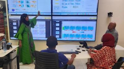 Four people in an office look at large screens displaying maps and data charts; one woman stands and points to the screen while others sit and observe.