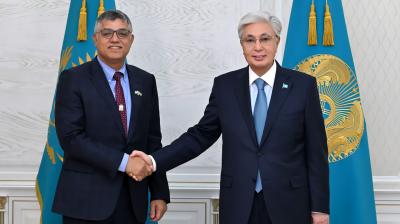 Two men in suits shake hands in front of two blue flags with gold emblems in a formal setting.