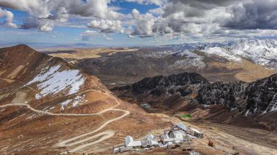 Mountain landscape with a winding road leading to buildings at the base, surrounded by rocky hills, snow patches, and distant snow-capped peaks under a cloudy sky.