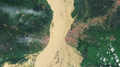 A wide, muddy river separates two densely built urban areas, surrounded by green vegetation and patches of cloud cover, as seen from above.
