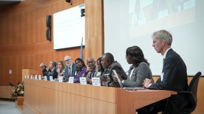A panel of speakers sits at a long table in a conference room, participating in a formal discussion with microphones and nameplates in front of them.