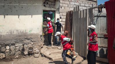 Red Cross workers in red vests walk toward a house with stone and brick walls; one person carries a child and another man stands by the door.