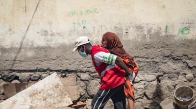 A man wearing a mask and red vest carries a woman on his back past a weathered wall and scattered debris.