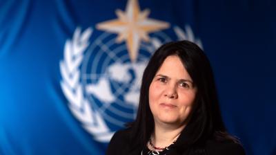 A woman with long dark hair in a black suit stands in front of a blue backdrop featuring a white emblem with a globe, laurel wreath, and a gold star.