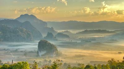 A scenic landscape with mountains, rolling hills, and mist covering the valley under a partly cloudy sky at sunrise.