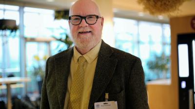Bald man with glasses, beard, and mustache wears a suit, yellow shirt, and tie, standing indoors with a conference name badge clipped to his jacket.