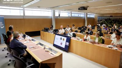 People attend a conference in a modern meeting room, with speakers at a front panel and a large screen displaying a presentation; GEO Group on Earth Observations logo is visible.