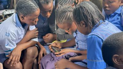 A group of schoolgirls in uniforms sit closely together, looking intently at a smartphone, inside a classroom.