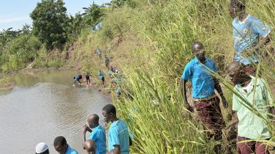 A group of students in blue uniforms walk along a grassy riverbank, with some standing near the water and others climbing the slope.