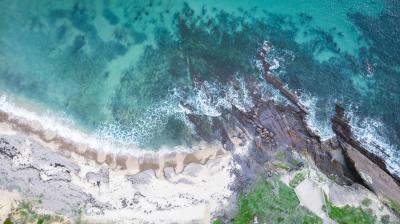Aerial view of a rocky shoreline with waves crashing onto a sandy beach and clear turquoise water.