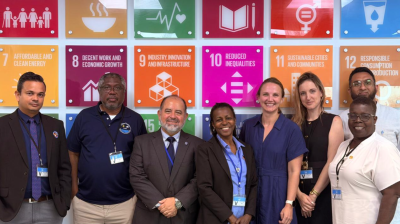 Eight people stand smiling in front of a colorful wall displaying icons and goals related to the United Nations Sustainable Development Goals.