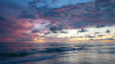 Waves gently roll onto a beach under a colorful sunset sky with shades of blue, pink, and purple, scattered with clouds.