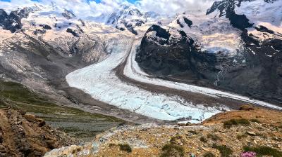 A winding glacier flows between rocky, snow-capped mountains under a partly cloudy blue sky, with sparse vegetation in the foreground.
