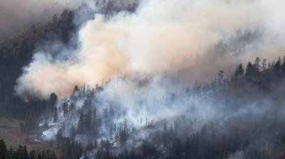 A wildfire burns through a forested hillside, producing thick smoke that rises and spreads across the landscape.