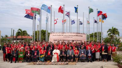 A large group of people pose for a photo in front of international flags and a banner for the Pacific Meteorological meeting, with palm trees and a clear sky in the background.