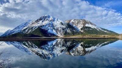 Snow-capped mountains are reflected in a calm lake under a blue sky with scattered clouds.